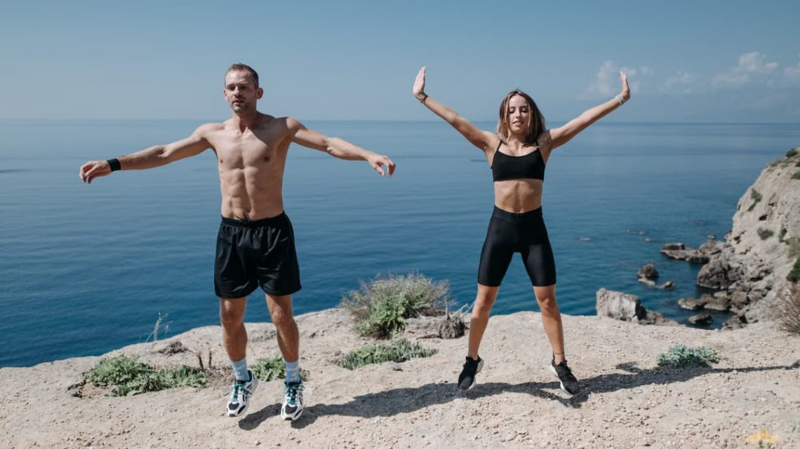 A man and woman perform jumping jacks on a cliff by the ocean under a clear sky.