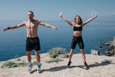 A man and woman perform jumping jacks on a cliff by the ocean under a clear sky.