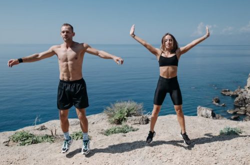 A man and woman perform jumping jacks on a cliff by the ocean under a clear sky.