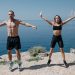 A man and woman perform jumping jacks on a cliff by the ocean under a clear sky.