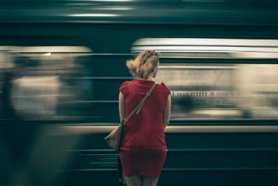 A woman in a red dress waits as a subway train speeds by in a blur, creating a sense of motion.