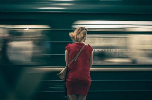 A woman in a red dress waits as a subway train speeds by in a blur, creating a sense of motion.