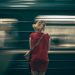 A woman in a red dress waits as a subway train speeds by in a blur, creating a sense of motion.